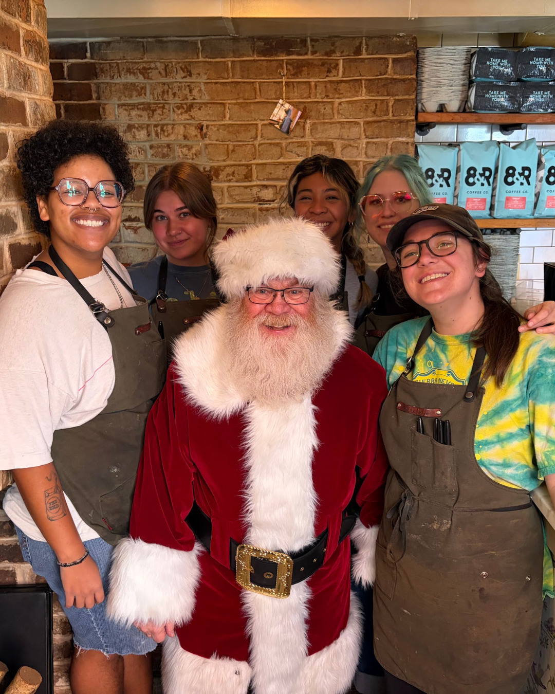 Group of five women and a man dressed as Santa Claus, smiling in a cozy coffee shop with a brick wall background.
