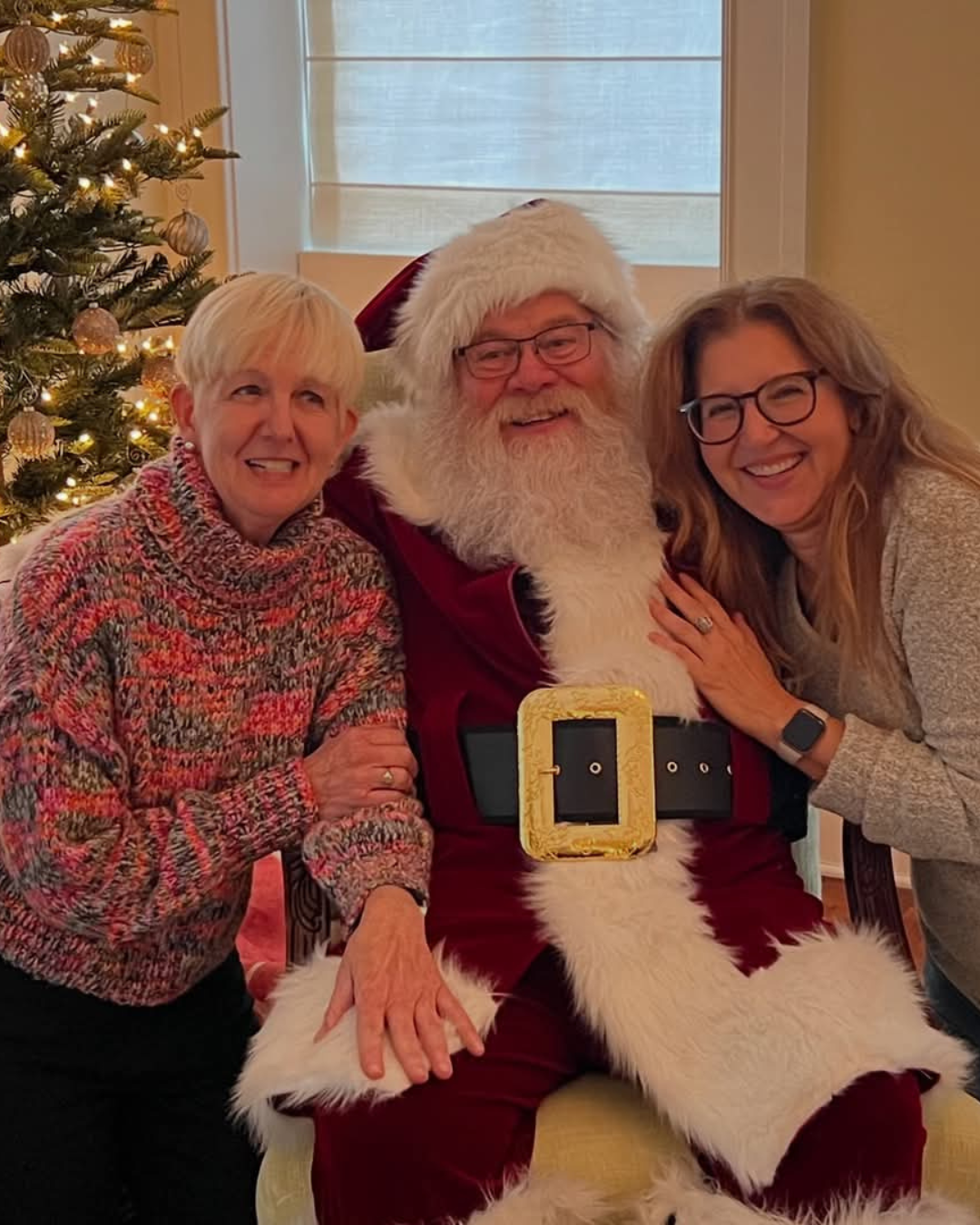 Two women and Santa Claus sitting near a decorated Christmas tree, smiling for the photo. One woman has short blonde hair and is wearing a multicolored sweater, the other has long blonde hair and glasses, wearing a gray sweater. Santa is dressed in t