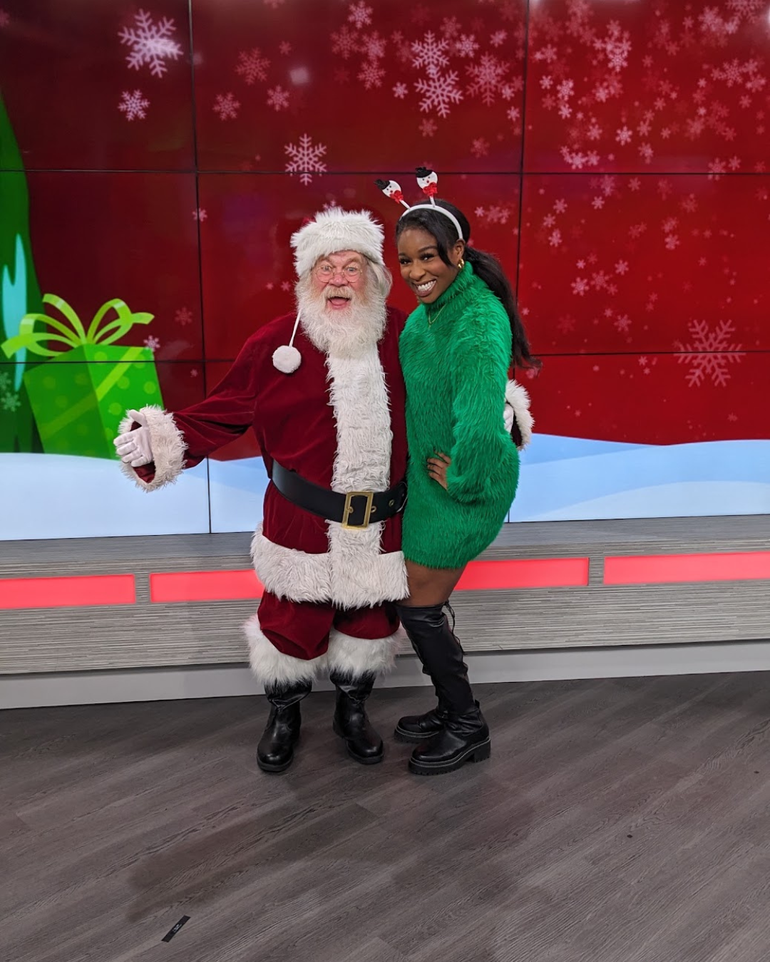 A woman in a green Christmas sweater dress, black thigh-high boots, and holiday headband with snowmen, posing with Santa Claus in a red and white costume, in front of a festive background with snowflakes and a green gift box.