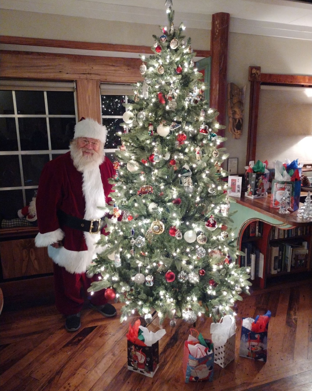 Santa Claus standing next to a decorated Christmas tree in a cozy living room, surrounded by gift bags and holiday decorations.