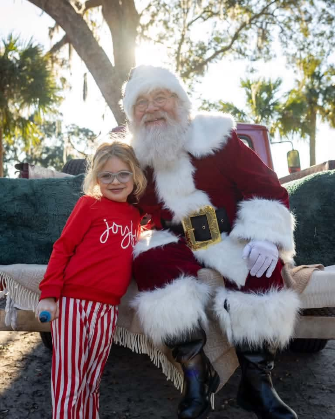 A young girl in red pajamas with a joyful expression standing next to a man dressed as Santa Claus, who is sitting on a bench outdoors with sunlight filtering through trees.