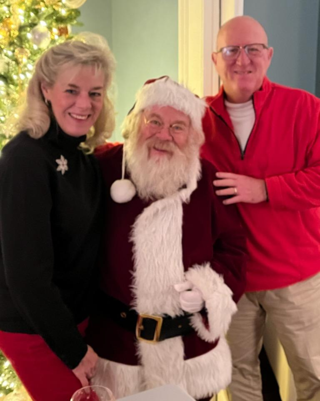 A woman and a man standing next to Santa Claus, who is dressed in a traditional red suit and hat, in front of a decorated Christmas tree.