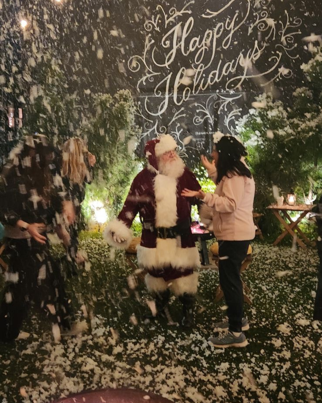 Santa Claus in a red suit and hat talking with a woman at a holiday event, surrounded by artificial snow and Christmas greenery, with "Happy Holidays" written on a black background above.