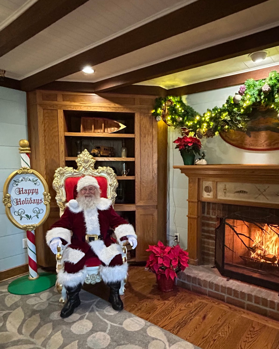 Santa Claus sitting on an ornate gold and red chair in front of a fireplace, decorated with a garland and Christmas ornaments. A sign next to Santa reads 'Happy Holidays'. Poinsettias are placed near the fireplace.