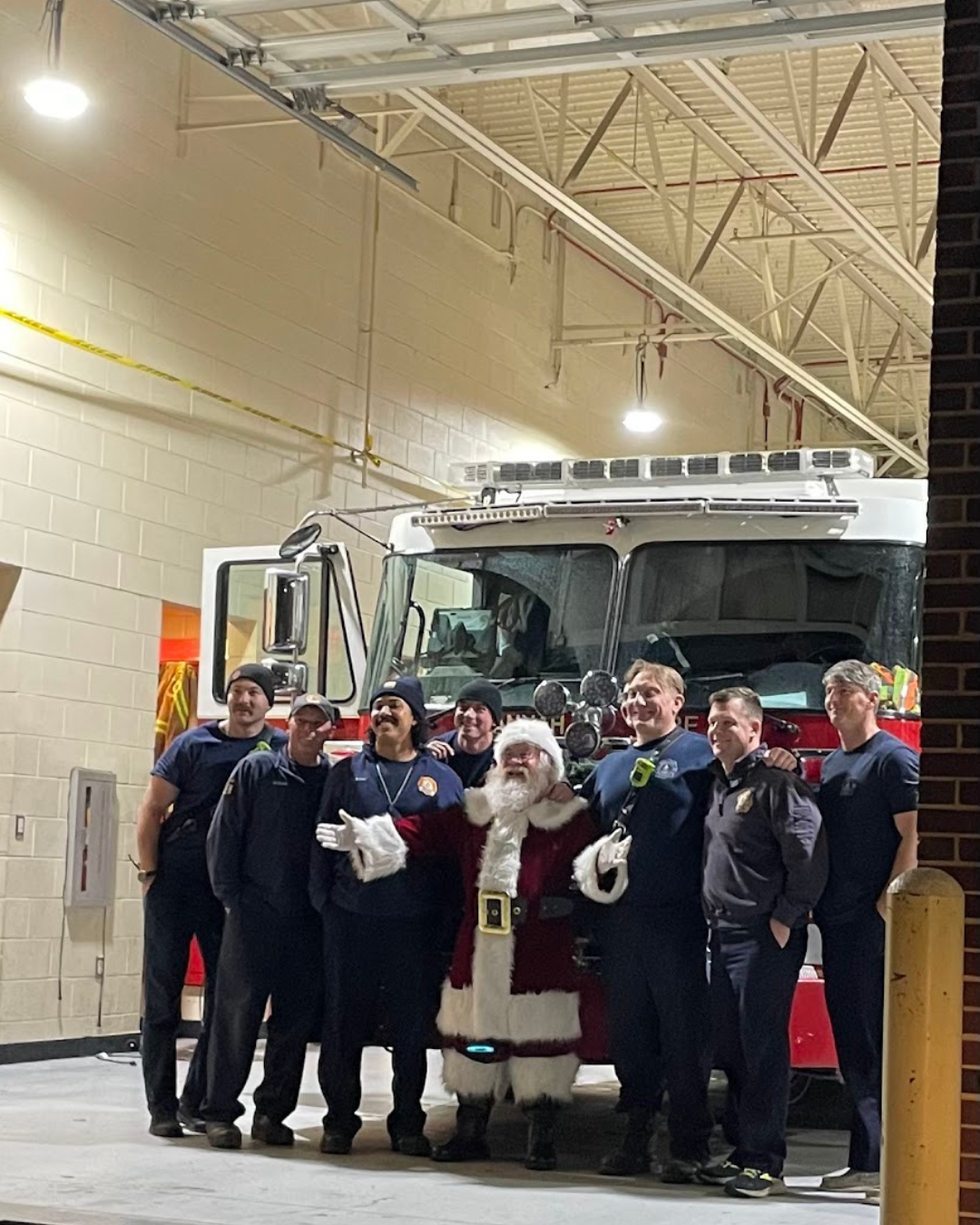 A group of firefighters, including one dressed as Santa Claus, posing in front of a fire truck inside a fire station garage.