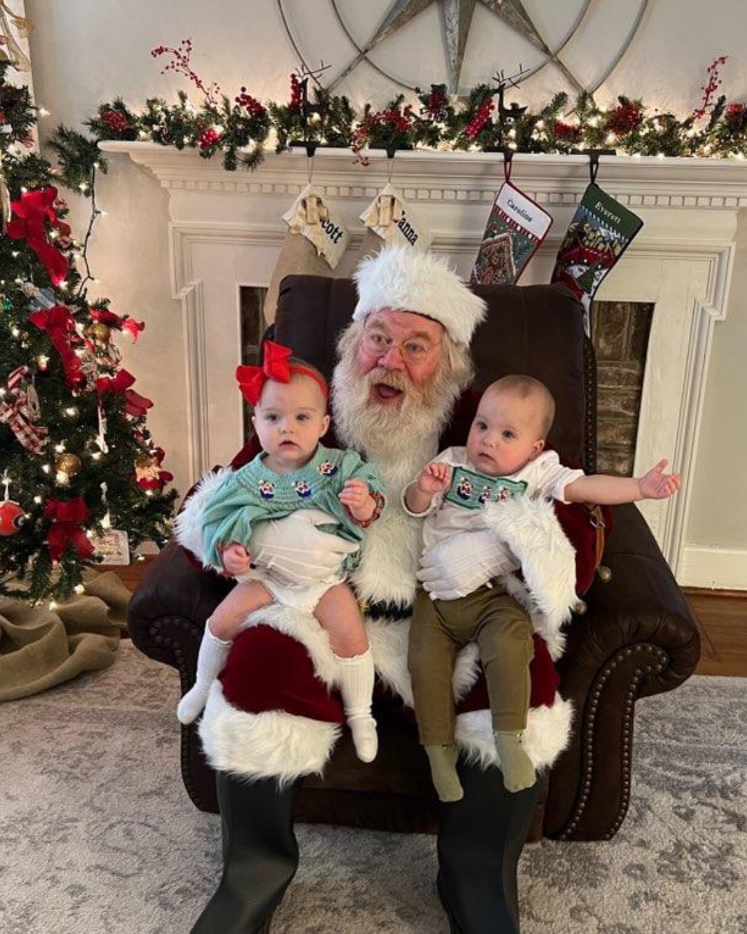 Santa Claus with two young children in front of a decorated Christmas fireplace, Christmas tree with ornaments, stockings hanging, and holiday garland.