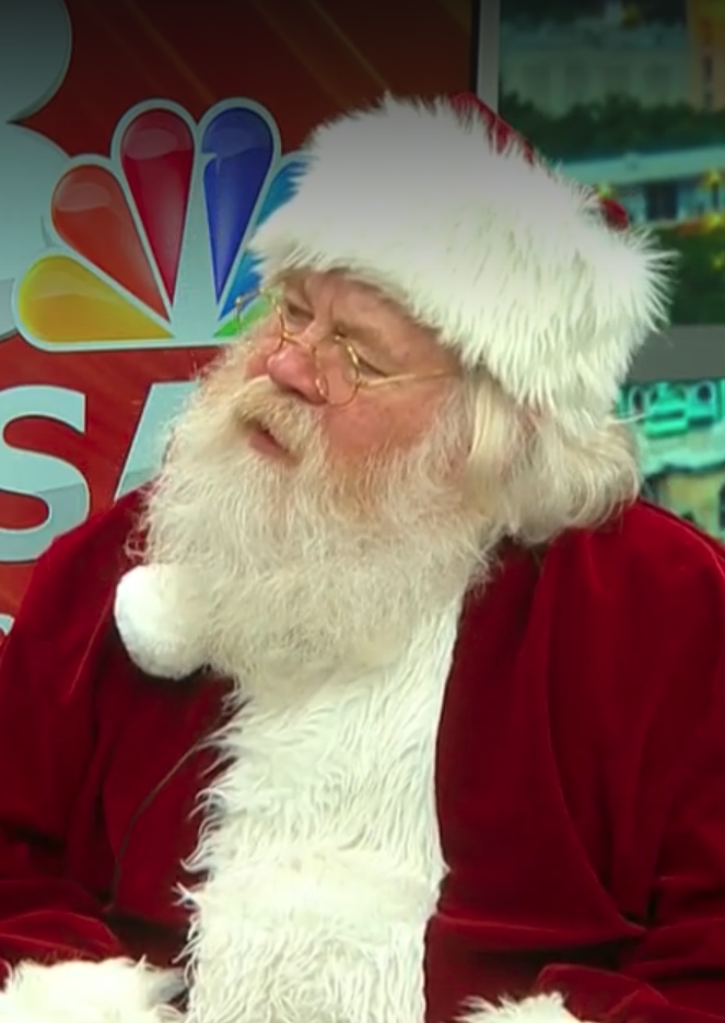 A man dressed as Santa Claus with a red suit, white beard, glasses, and a red and white Santa hat, sitting indoors with a NBC logo in the background.