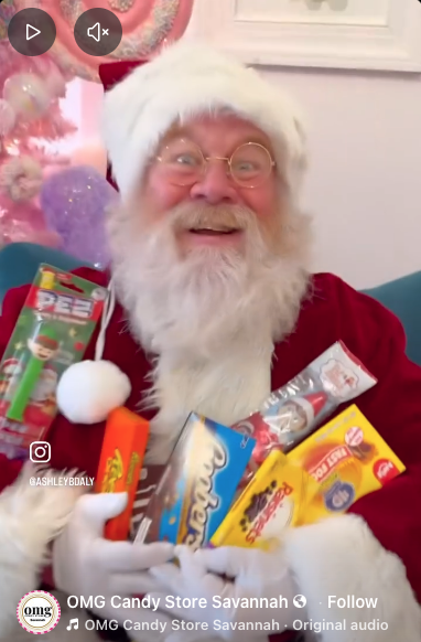 Santa Claus smiling and holding various Christmas candies and treats, with a decorated pink Christmas tree in the background.