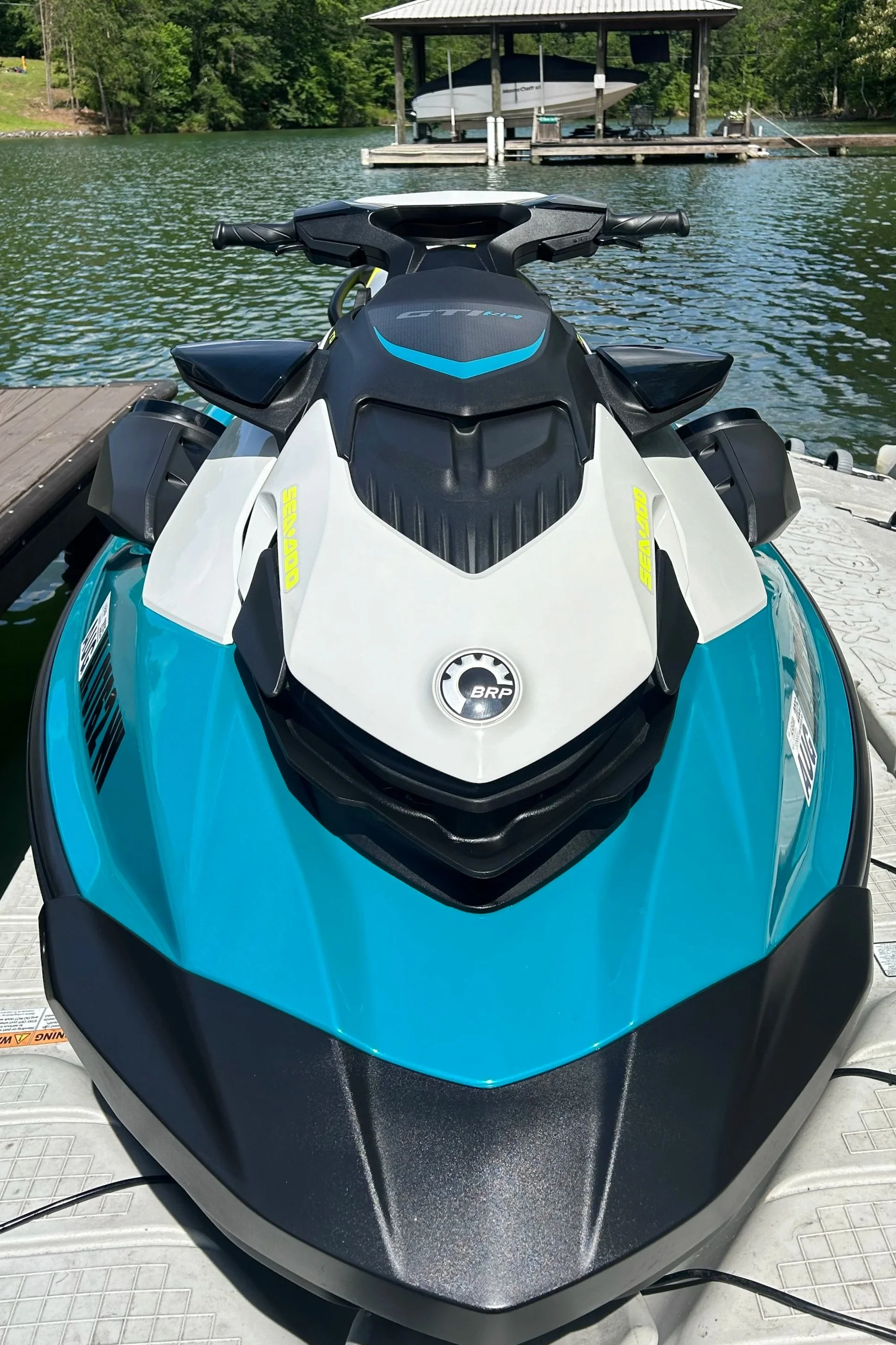 Front view of a teal and white BRP Sea-Doo personal watercraft docked at a lakeside with a boat lift and boat in the background.