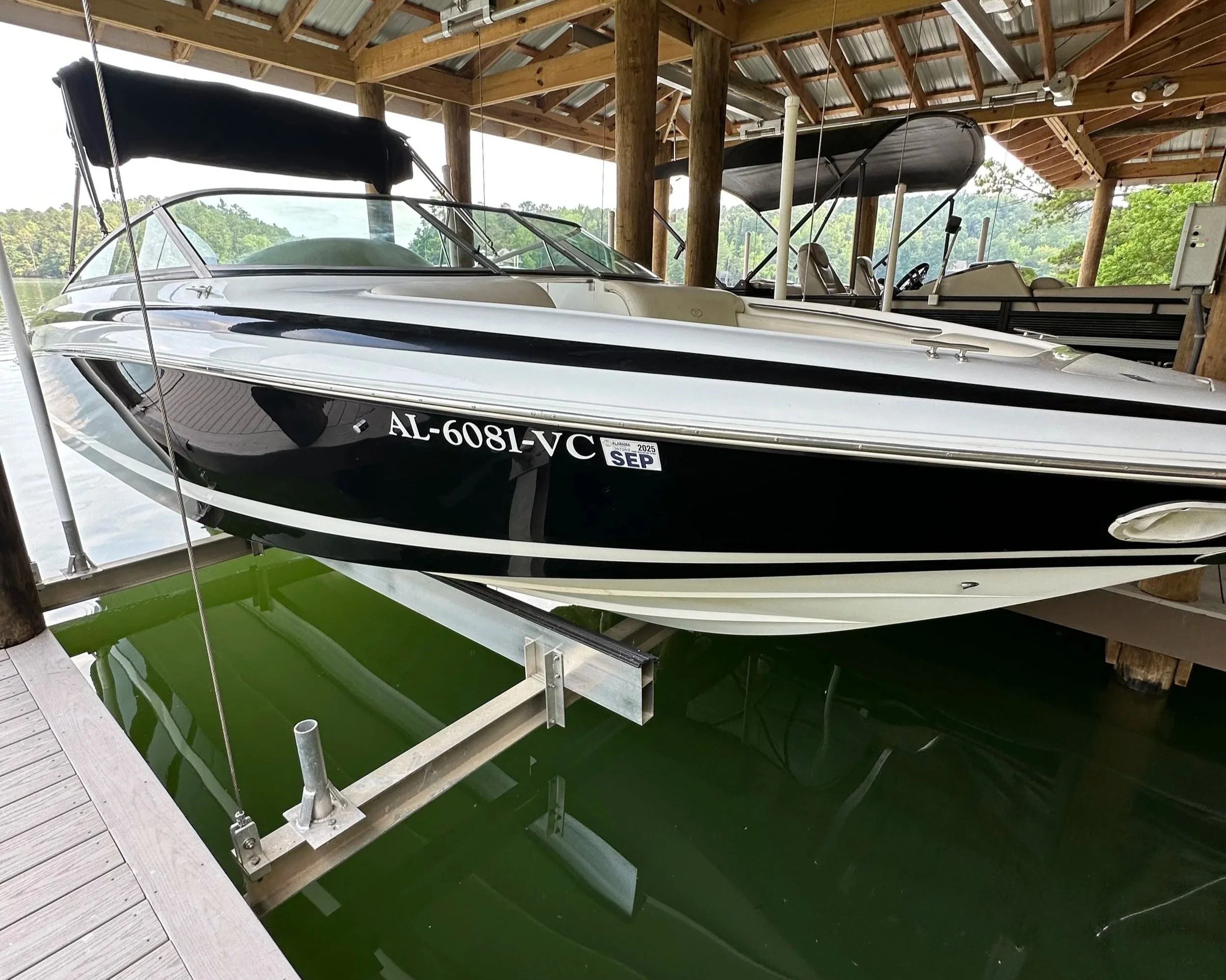 A black and white motorboat with registration AL-6081-VC docked inside a wooden boat shed on green water, with two other boats and trees visible in the background.