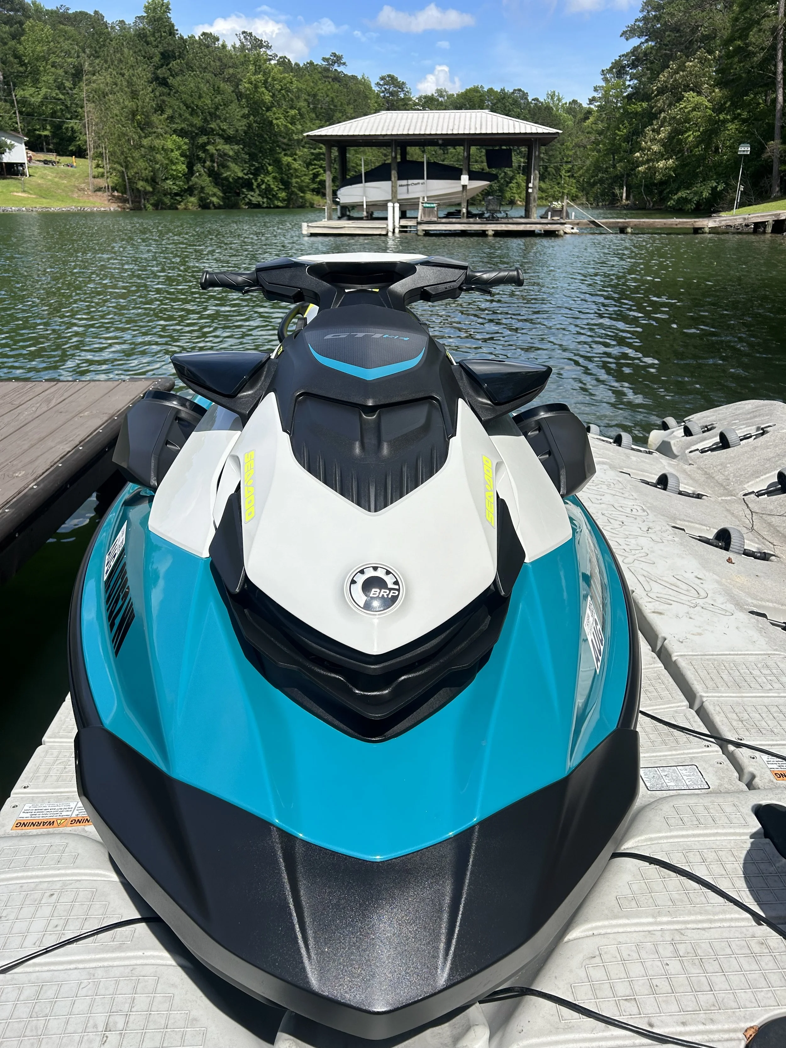 A blue and white jet ski docked at a boat ramp by a lake with a boat lift and dock in the background, surrounded by green trees.