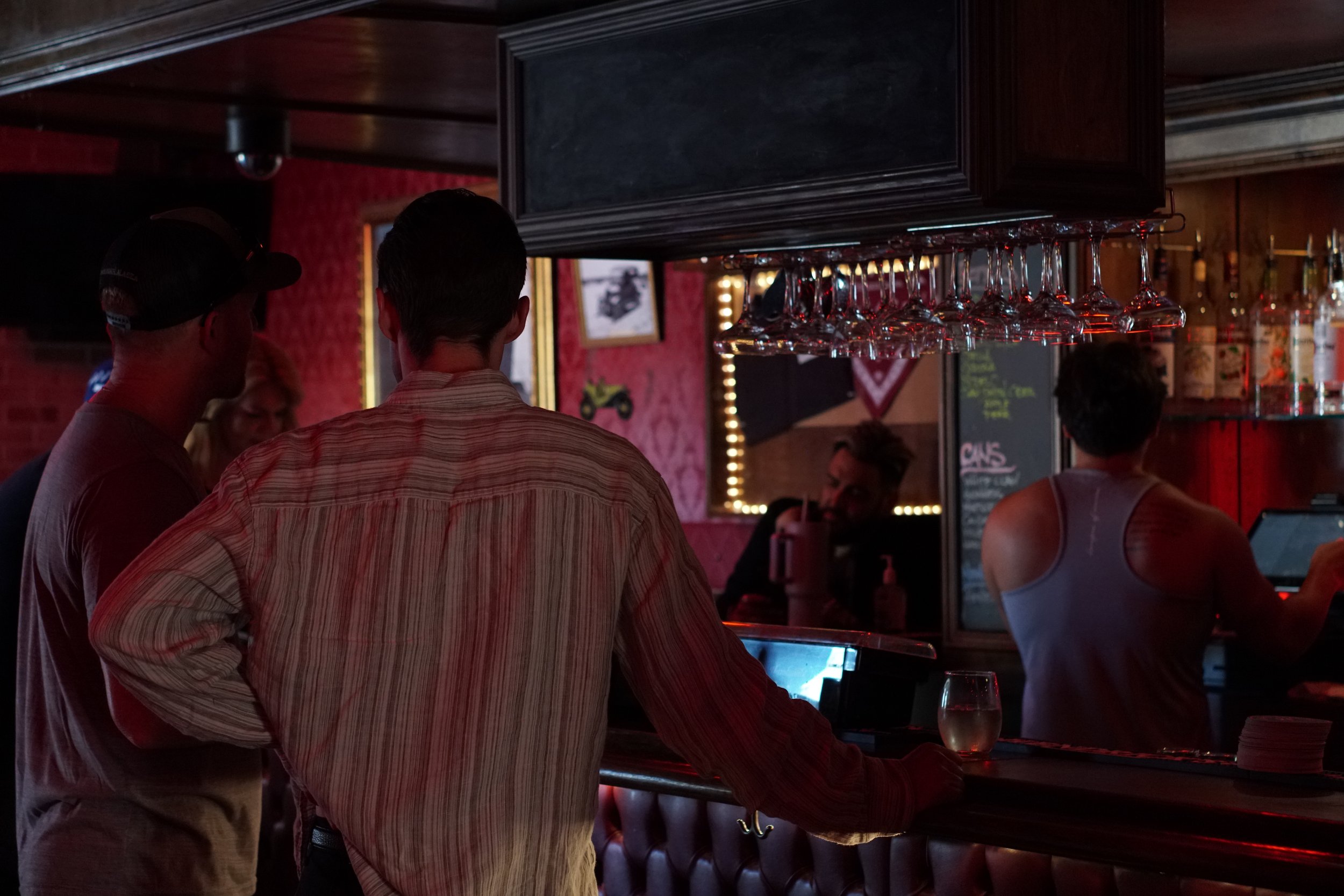 People at a dimly lit bar, with a bartender serving drinks and customers standing by the counter. Wine glasses hang upside down, and liquor bottles are visible in the background.