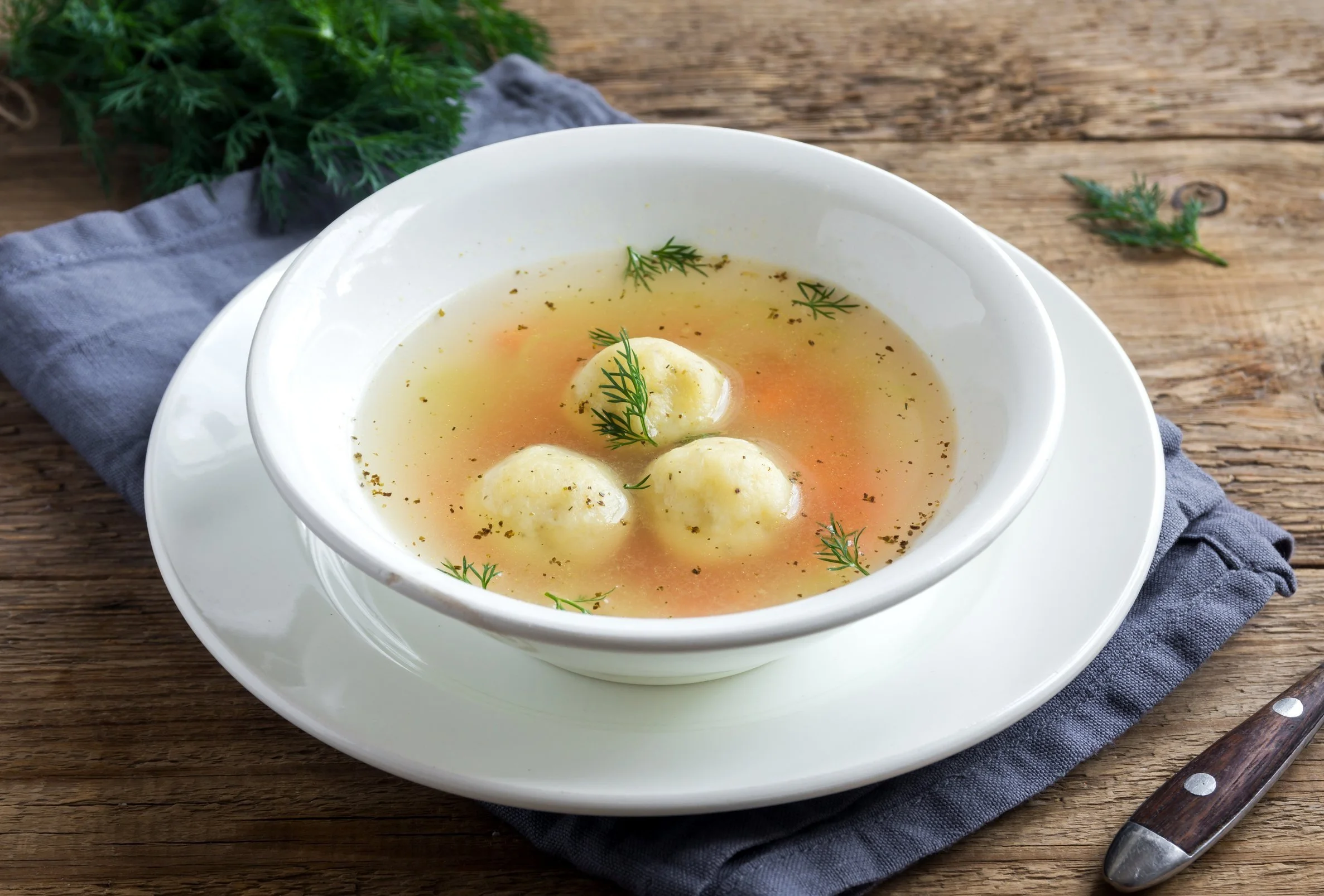 A bowl of soup with matzo balls, garnished with dill, on a plate with a napkin and a wooden table background.