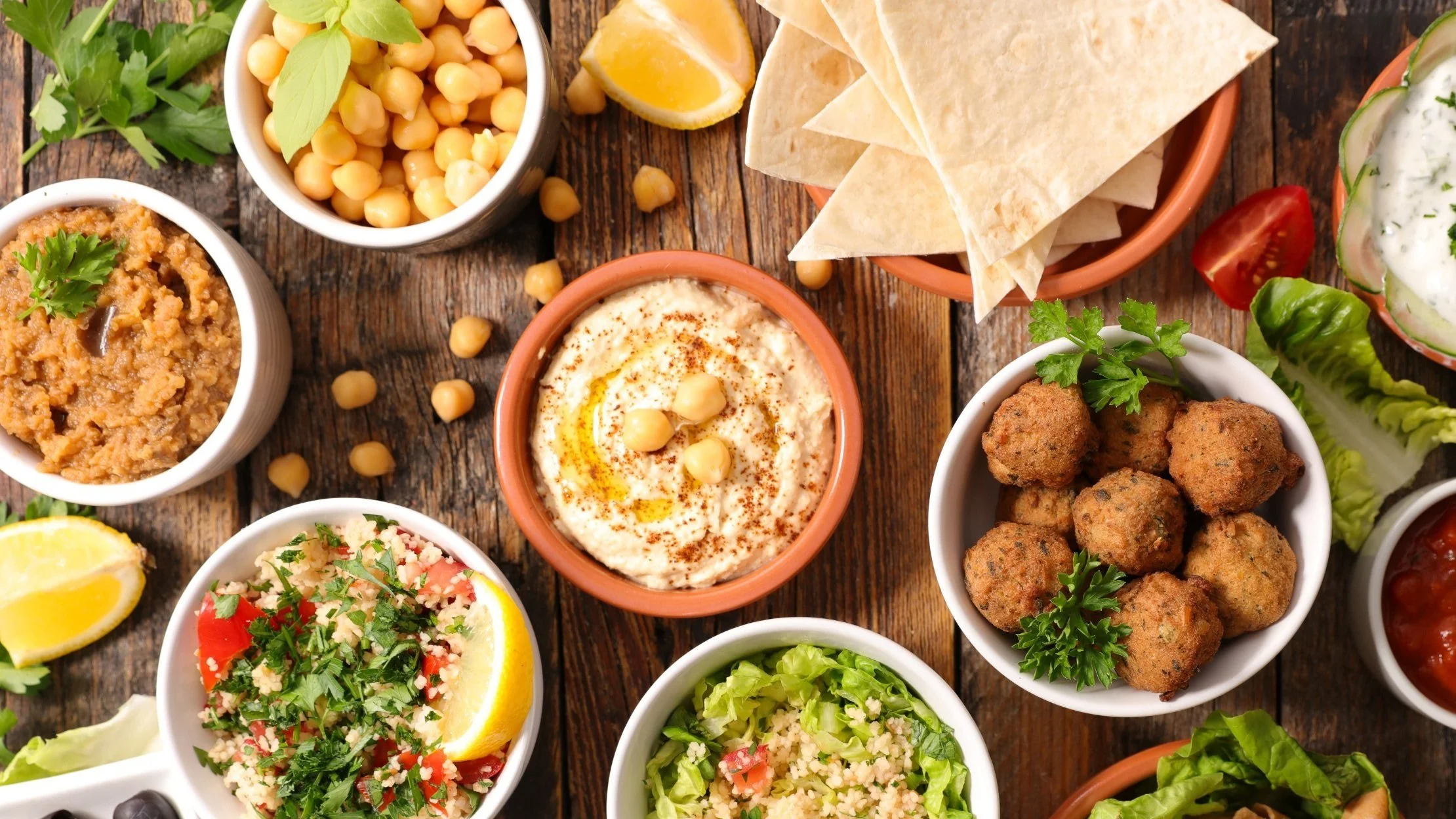 Assorted Mediterranean dishes including hummus, chickpeas, falafel, tabbouleh, pita bread, baba ghanoush, and vegetables on a wooden table.
