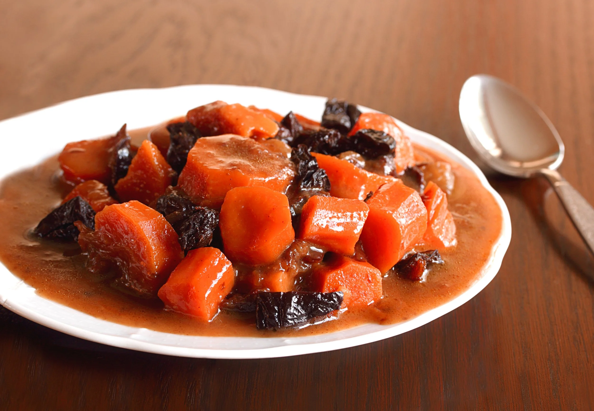 Tzimmes, a Jewish dish with carrots, prunes, and orange sauce on a white plate with a spoon.