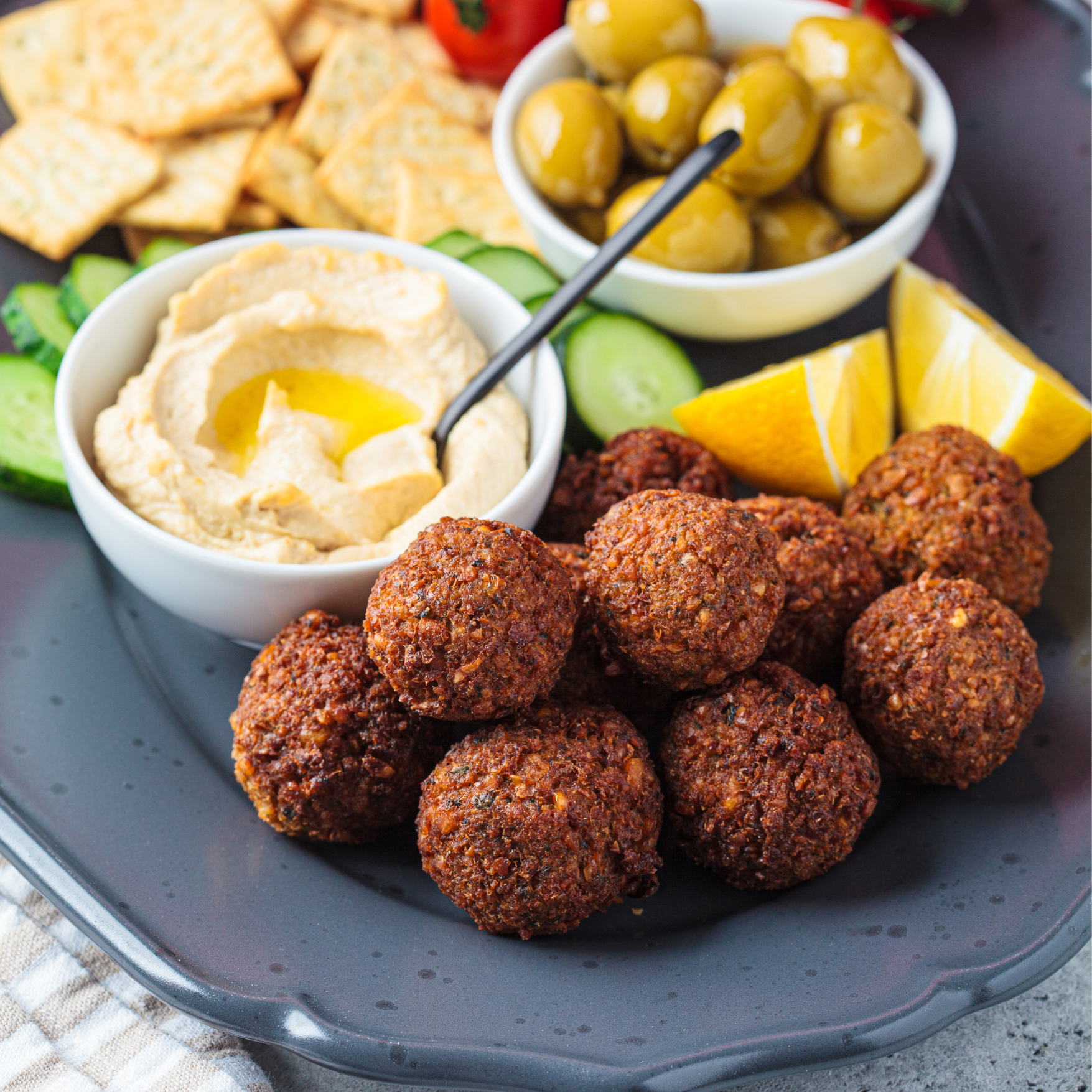 Plate with falafel balls, hummus, sliced cucumber, crackers, green olives, and lemon wedges.