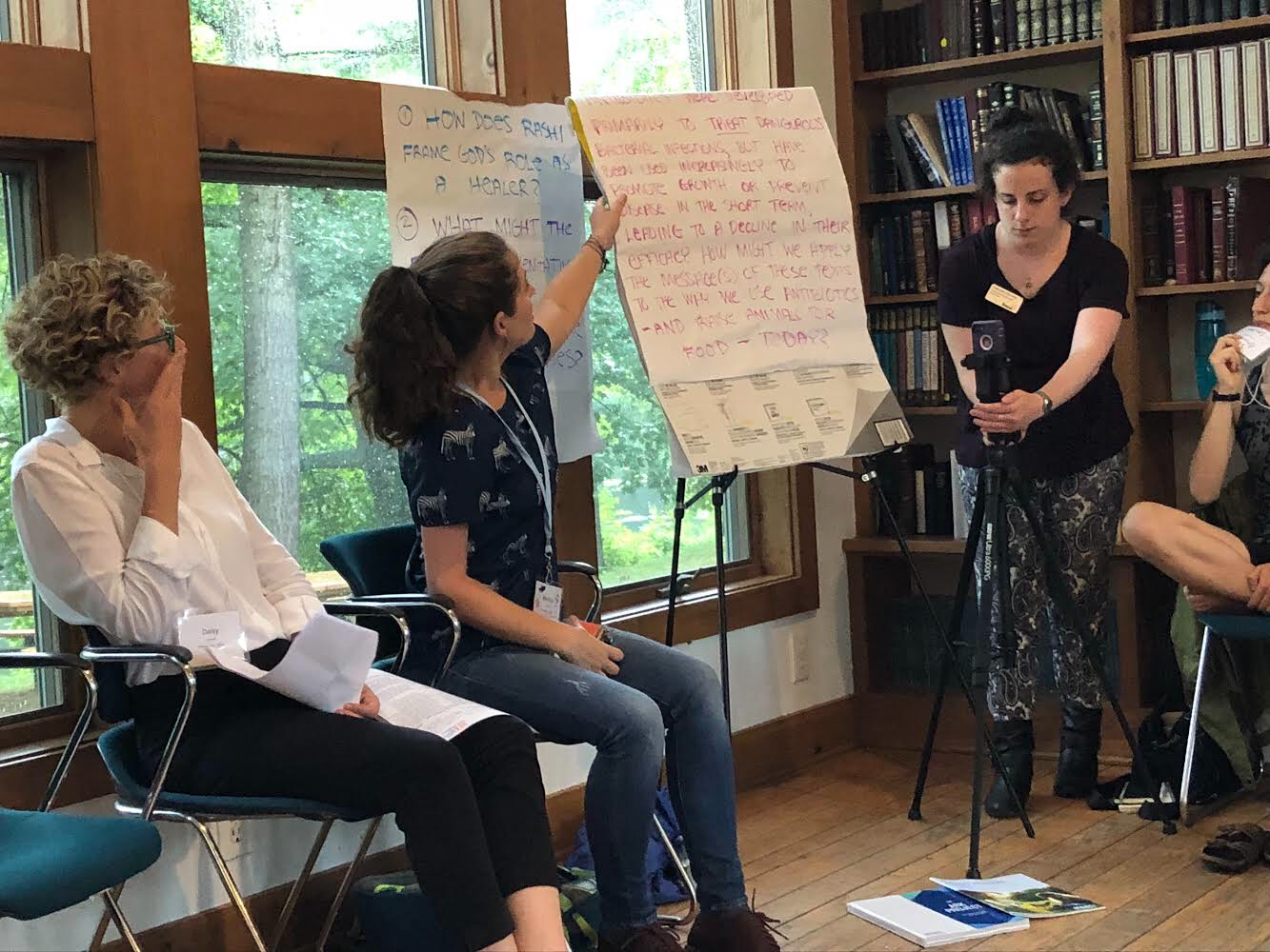 Group discussion in a library setting with three people, whiteboards, and a camera on a tripod.