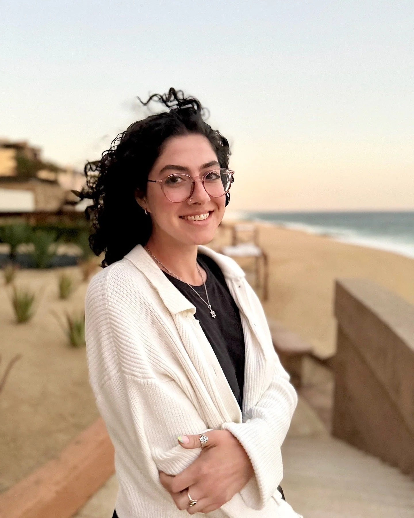 Person with curly hair smiling, wearing glasses and a white cardigan, standing on a beach with ocean in the background.