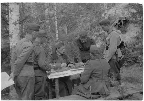 Group of soldiers seated and standing around a table outdoors, engaging in a discussion or briefing.
