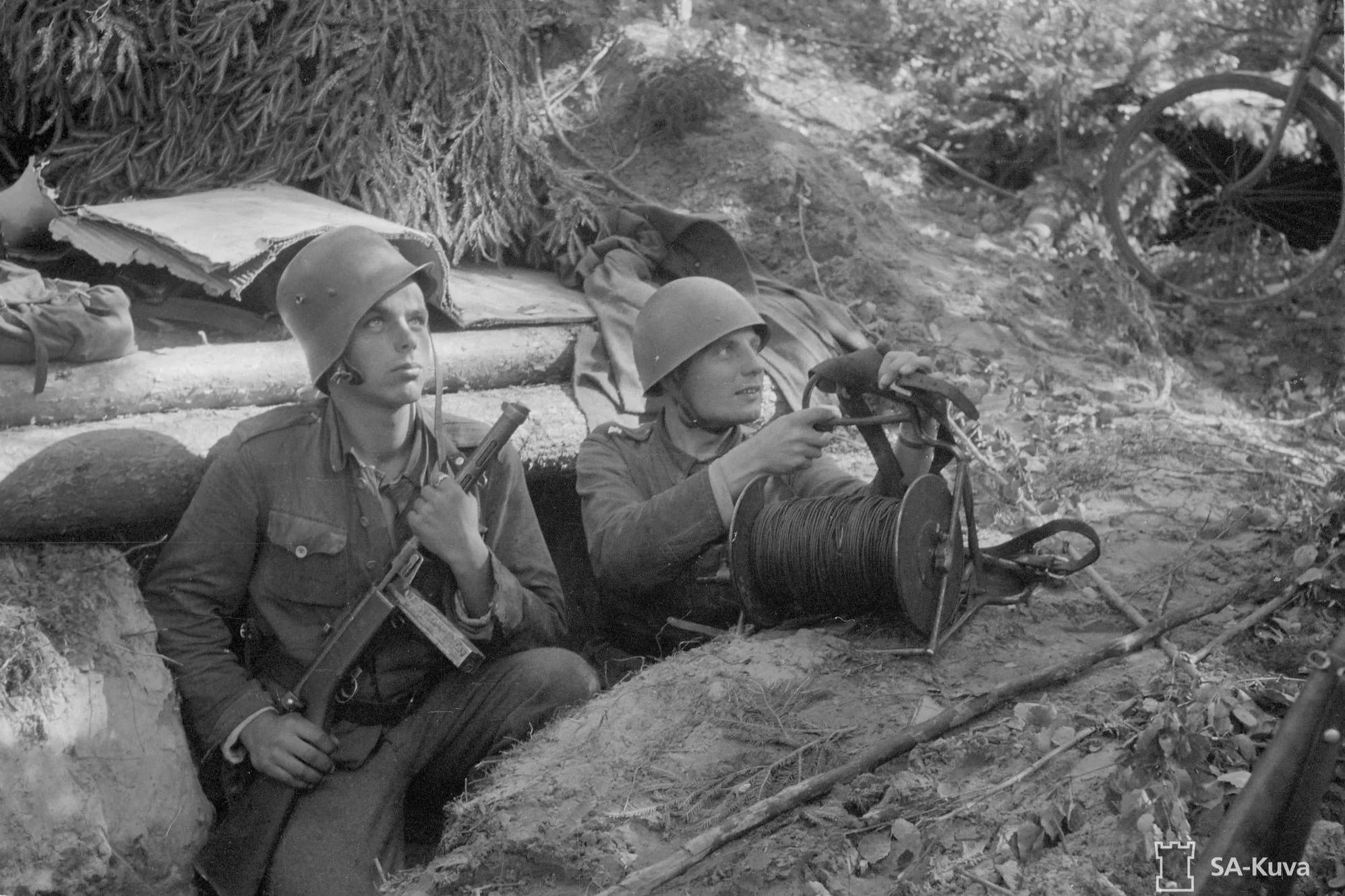 Two soldiers wearing helmets and military uniforms using a radio communication device in a natural outdoor setting.