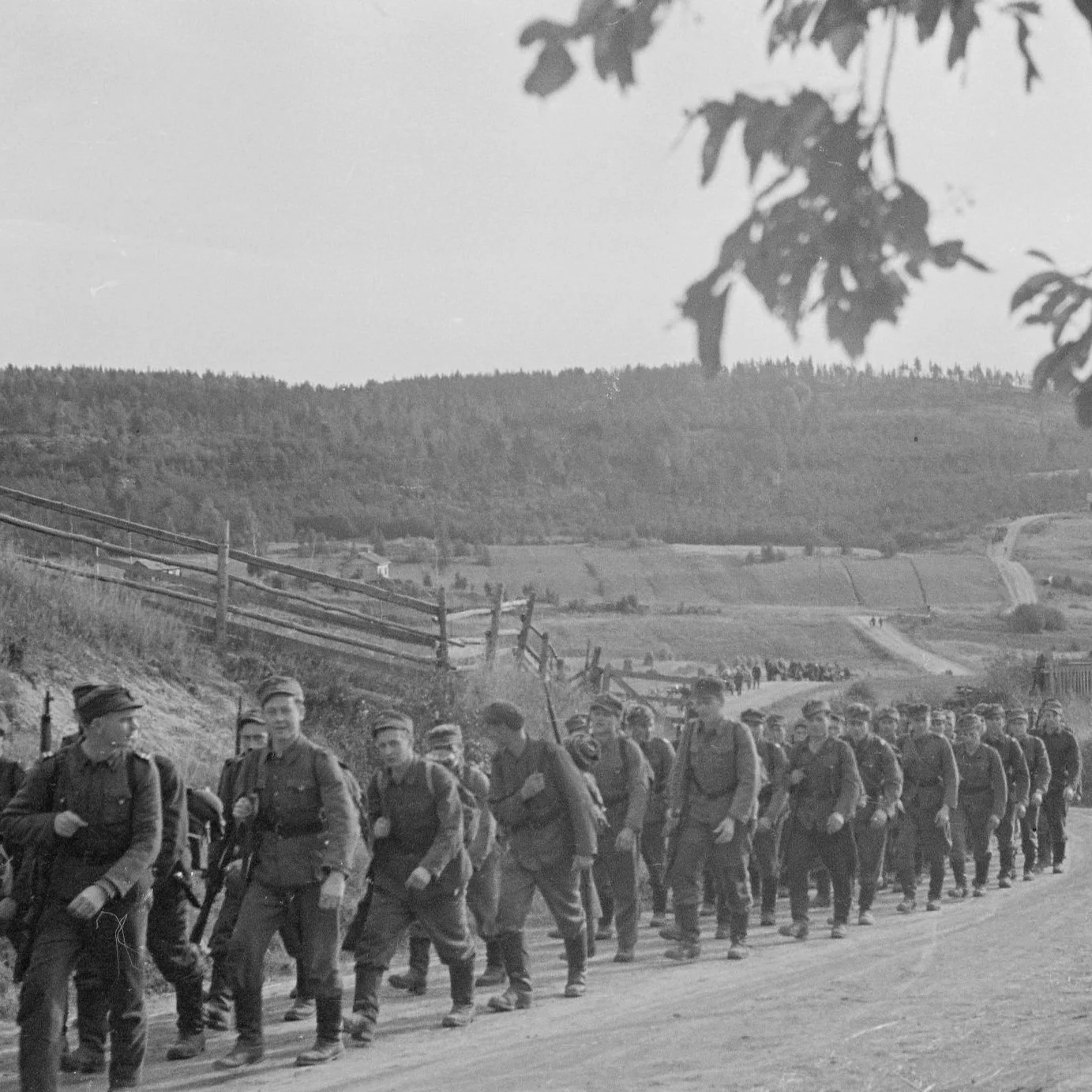 A group of soldiers marching on a rural road with hills and fields in the background in black and white.