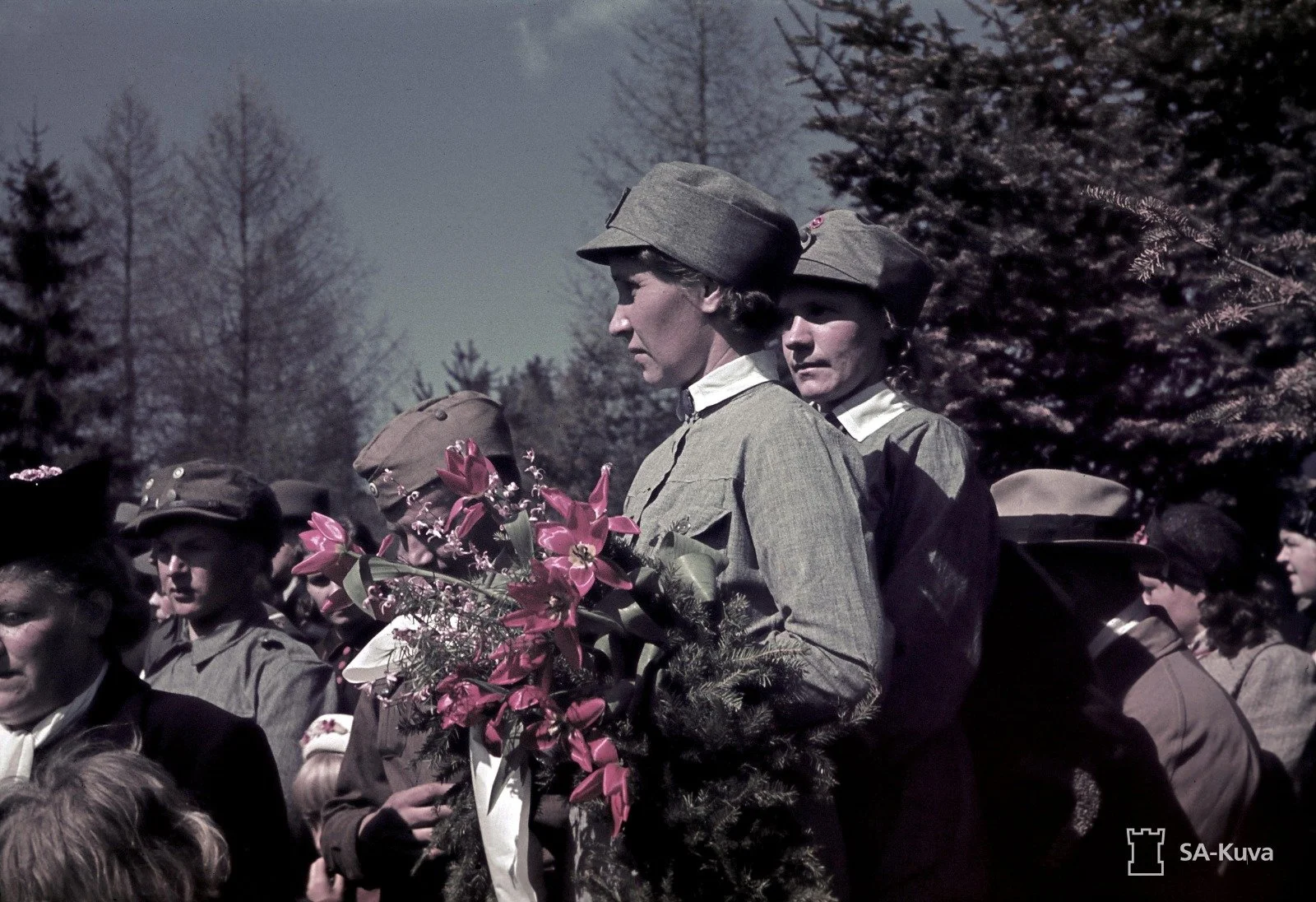 Women dressed in military uniforms participating in a memorial or commemorative event outdoors, with trees in the background, some women carrying flowers.