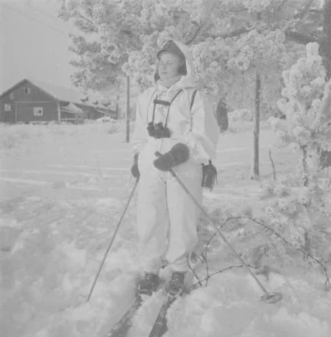 A person dressed in winter clothing with skis and a camera stands in a snowy landscape with trees and a building in the background.