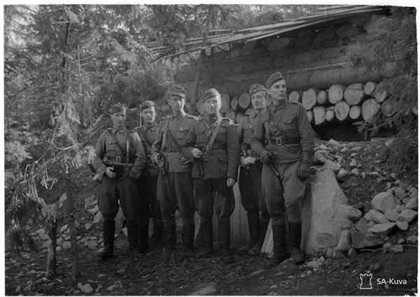 Black and white photograph of seven soldiers standing outdoors in front of a wooden shelter with logs stacked underneath. The soldiers are dressed in military uniforms and are holding rifles.