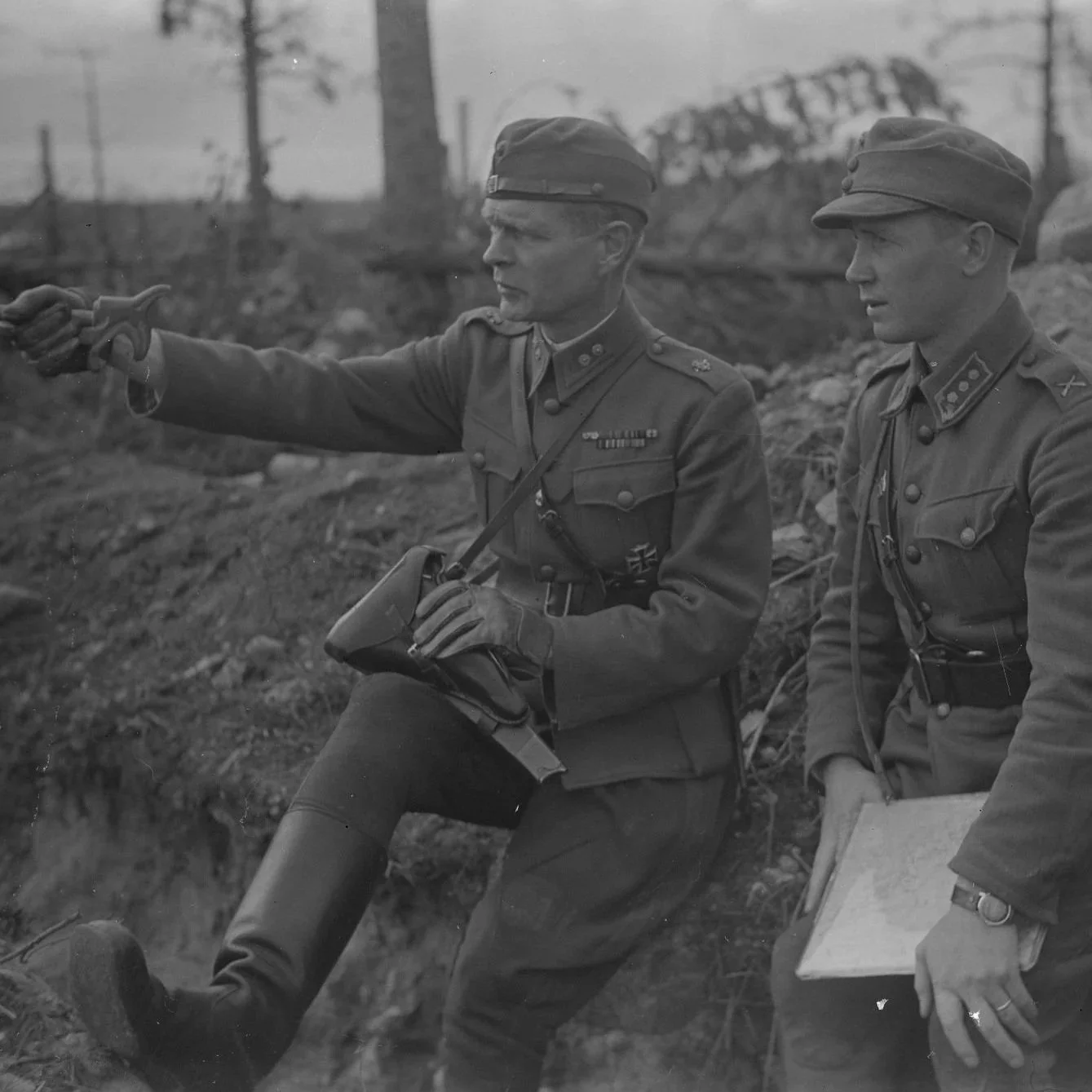 Two Finnish soldiers in military uniforms during wartime, one pointing with a Lynx shaped walking stick, and the other holding a map.