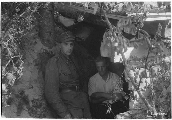Two men sitting under a tree near a cave or small shelter, with foliage surrounding them.