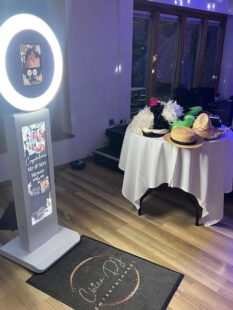 Photo of a wedding or event setup featuring a photo booth with a lighted circular frame, a sign that reads 'Congratulations Mr & Mrs Mower,' and a table with floral arrangements, hats, and miscellaneous items in front of large windows.