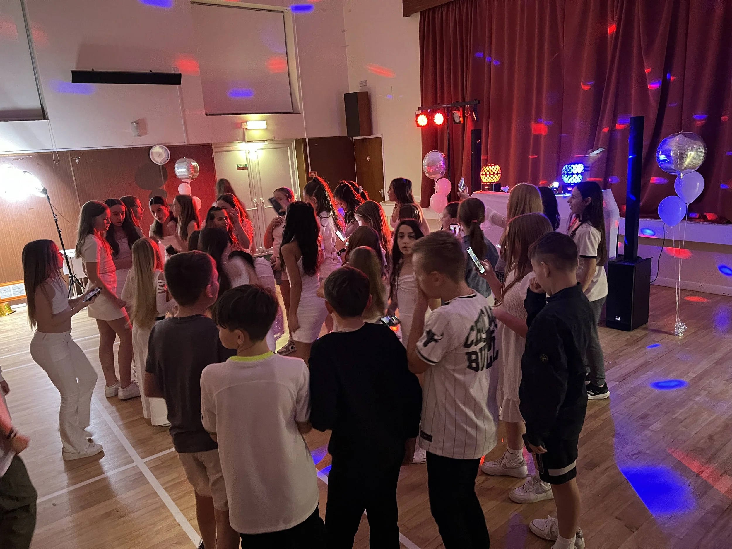 Group of children dancing and socializing at a party with colorful lights and balloons in a decorated hall.