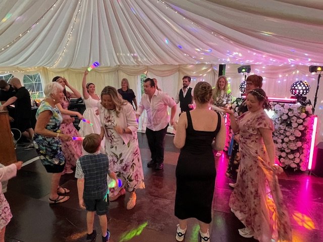 People dancing and enjoying music at a celebration under a decorated tent with draped fabric and colorful lights.