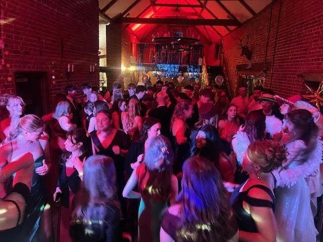 Crowd of people dancing and socializing in a dimly lit room with red lighting and exposed brick walls, possibly at a party or dance event.