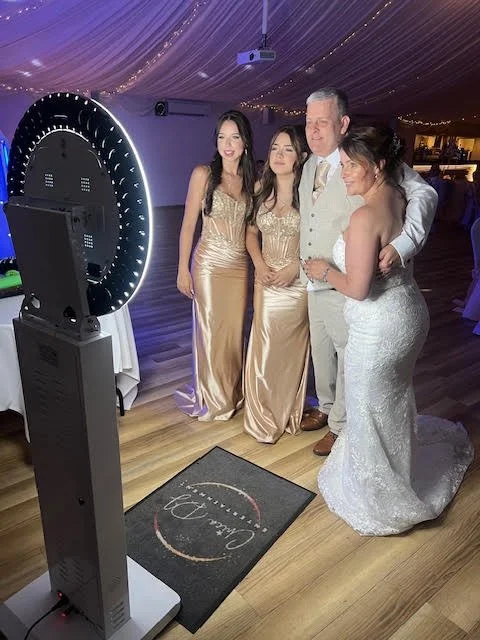 A bride and groom with three women posing in front of a photo booth at a wedding reception, with purple drapery and string lights in the background.
