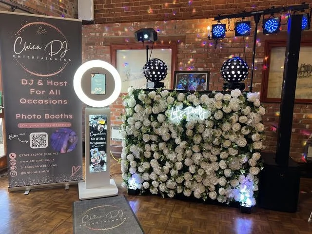 Event setup with a floral DJ booth decorated with white roses, colorful string lights, and professional sound equipment. There is a photo booth station on the left and a sign for Chica DJ Entertainment promoting DJ services for events.