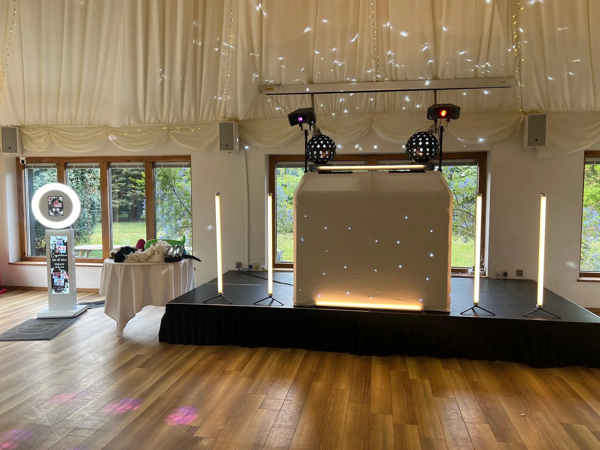 Event space with a stage, decorated with white drapes and string lights, with photo booth props and a shiny, illuminated backdrop for photos.