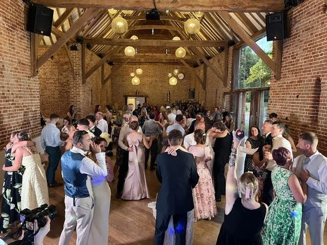Wedding reception with guests dancing in a rustic hall with brick walls, wooden beams, and hanging paper lanterns.