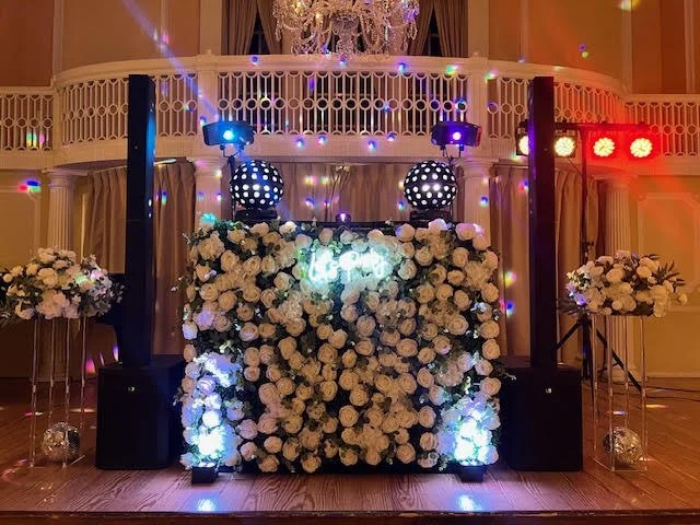 Event stage decorated with a large floral arrangement, spotlights, and colorful lighting in an elegant room with a balcony and chandelier.
