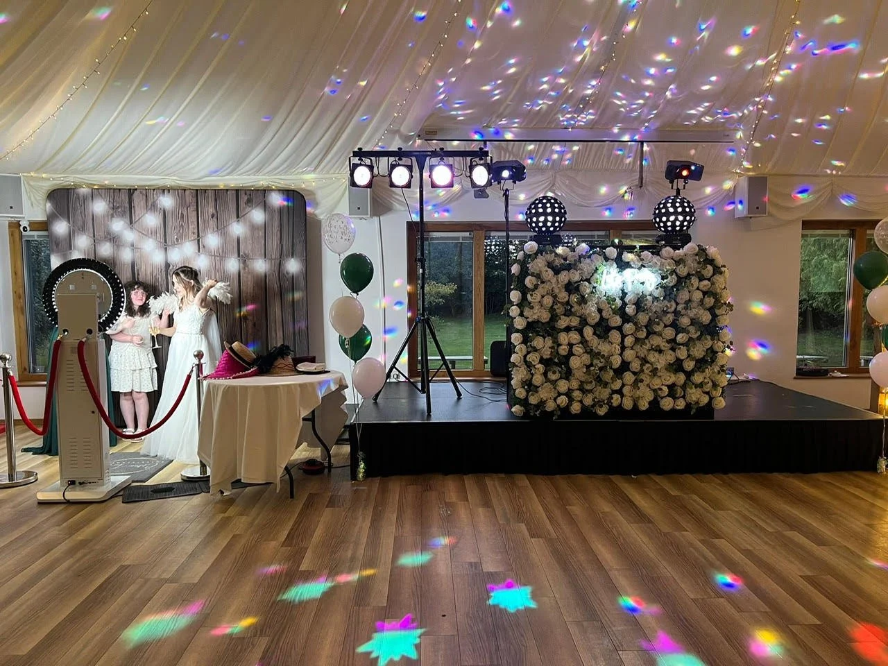 Decorated event space with a stage covered in white roses, lighting, and balloons, set up for a celebration with dancing. Two women are taking photos near a photo booth, and a table with props is nearby. Colorful lights create star-shaped patterns on