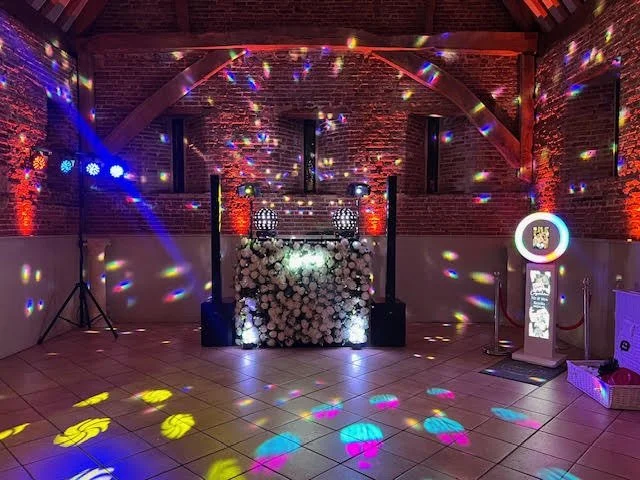 Empty indoor event space with disco lights, DJ booth decorated with white flowers, and colorful reflections on walls and floor.