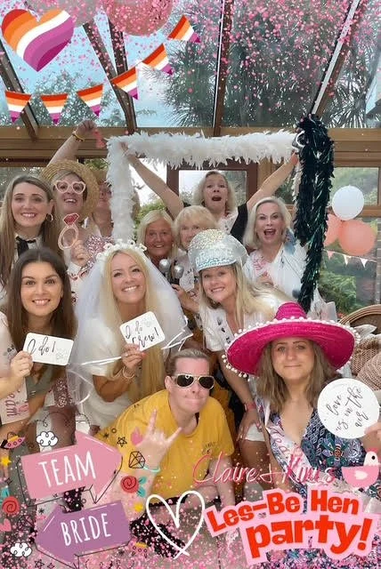 Group of women and girls at a hen party celebration, wearing festive accessories including hats, sunglasses, and holding signs, with colorful decorations and balloons.