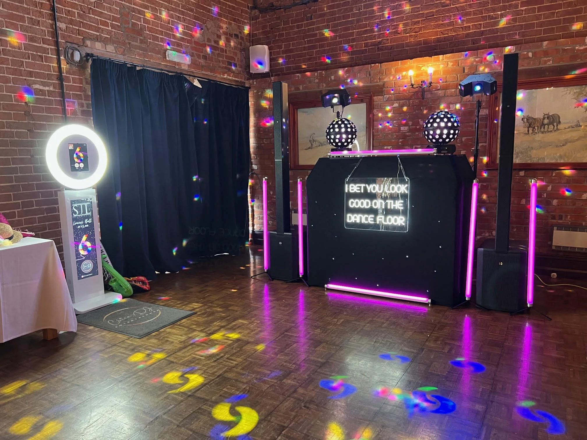 Dance floor with colorful disco lighting, two large spherical light fixtures, and a neon sign that reads 'I BET YOU LOOK GOOD ON THE DANCE FLOOR'. There is also a curtain, wall art, and a small table with a hat to the left.
