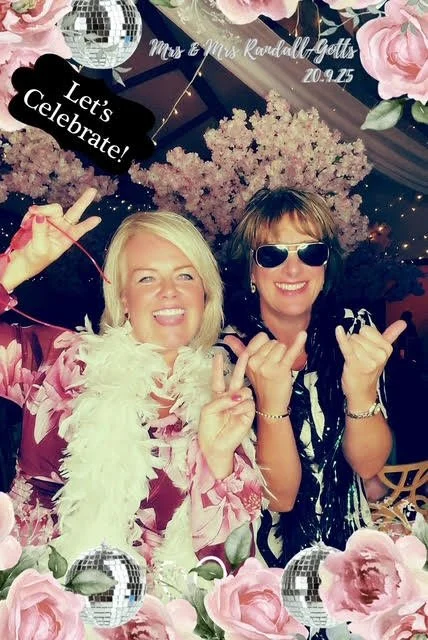 Two women smiling and celebrating at a party with pink flowers and decorative disco balls, one wearing sunglasses and the other making a peace sign, with a banner reading "Let's Celebrate" and a sign for Mrs. & Mrs. Randall Gotts, dated 7.25.2023.