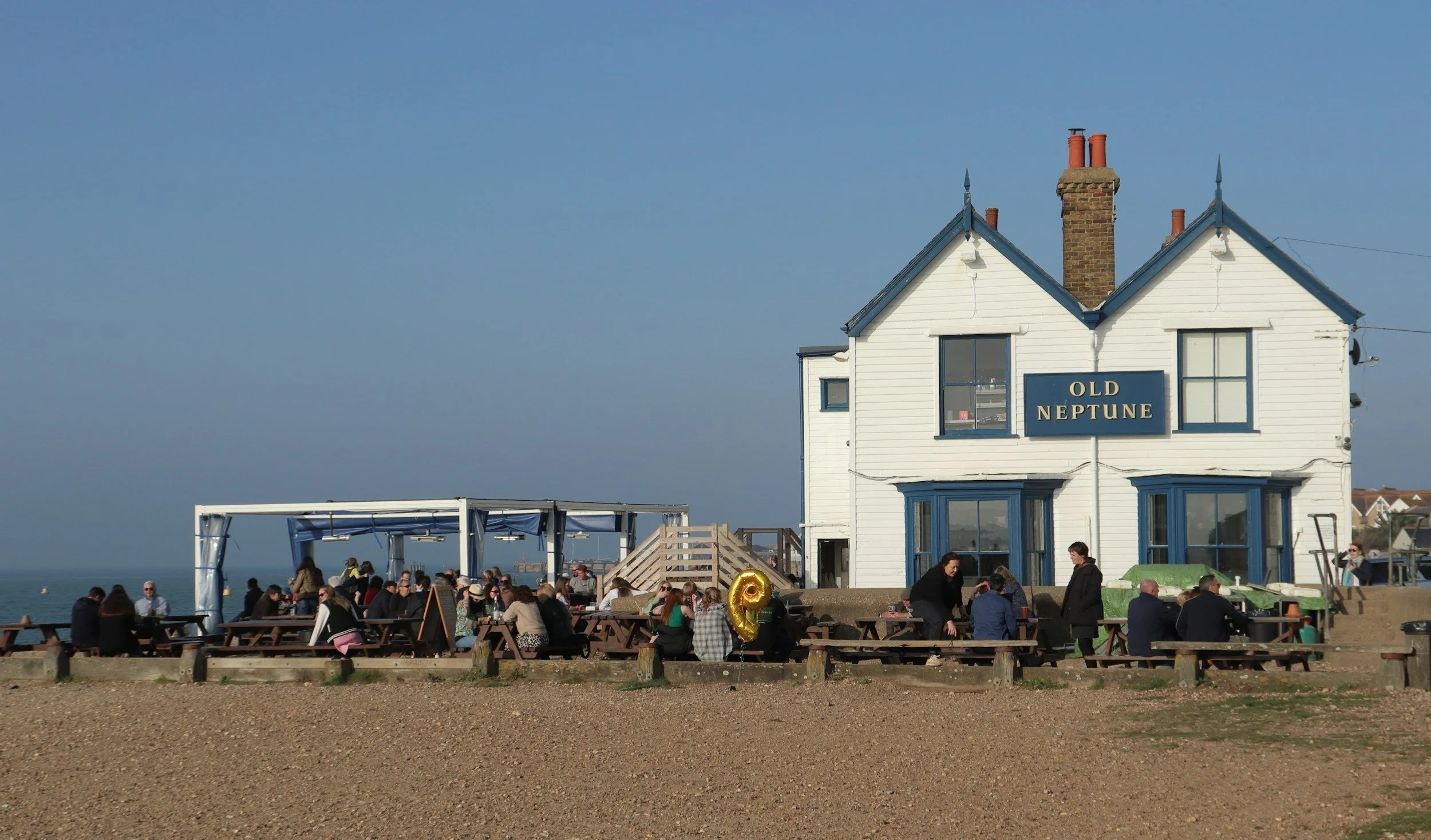 A lively outdoor restaurant scene with people dining at picnic tables in front of a white two-story building labeled 'Old Neptune,' by the seaside on a sunny day.