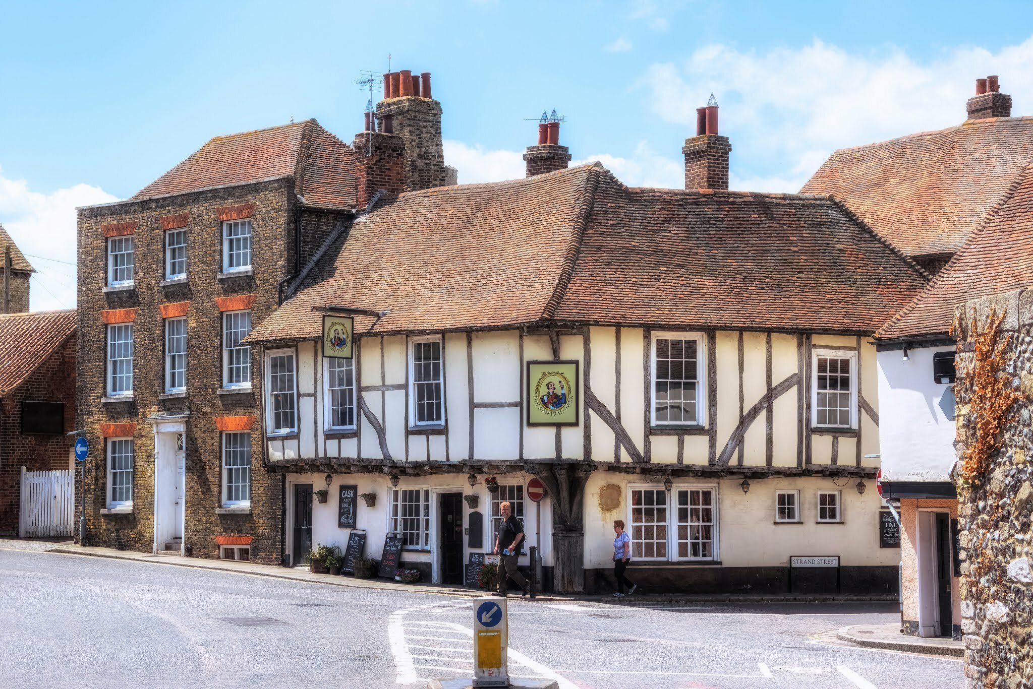 A street view of historic buildings including a traditional timber-framed pub with signs and outdoor blackboards, pedestrians walking, and a traffic island with a blue arrow sign under a partly cloudy sky.