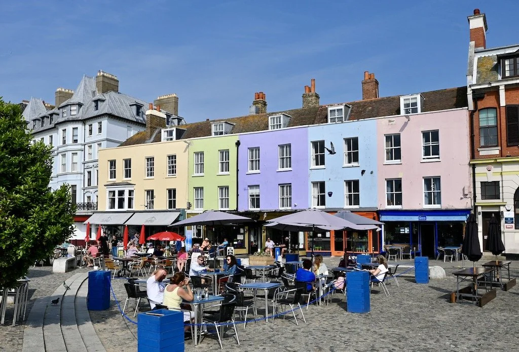 Colorful pastel buildings with outdoor cafe seating in a plaza, people dining and socializing under umbrellas, clear blue sky.