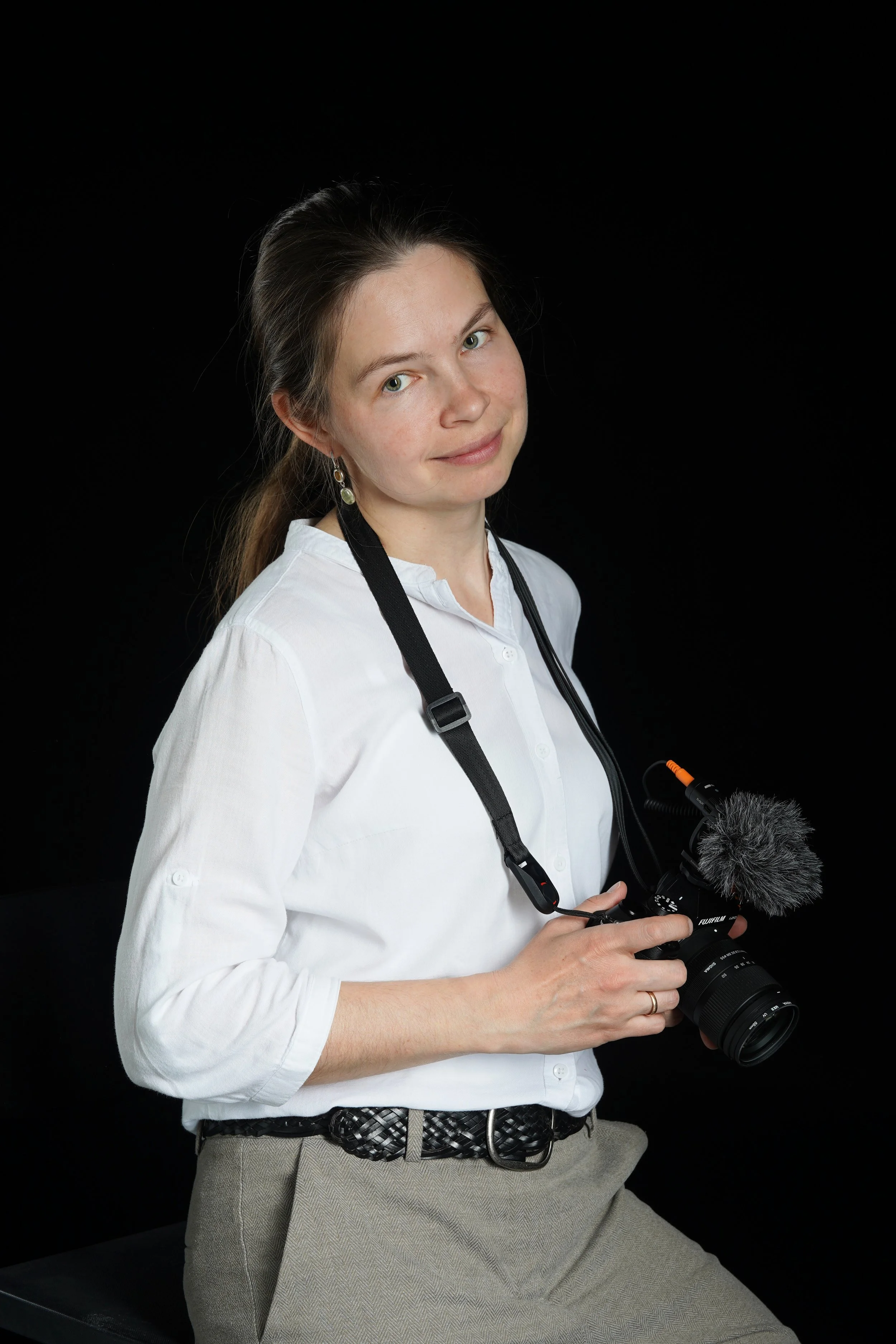 Eine Frau mit braunen Haaren, in weißem Hemd und hellgrauen Hosen, hält eine Kamera mit einem Mikrofoneinschub vor sich. Sie sitzt vor schwarzem Hintergrund und schaut leicht zur Seite, lächelnd.