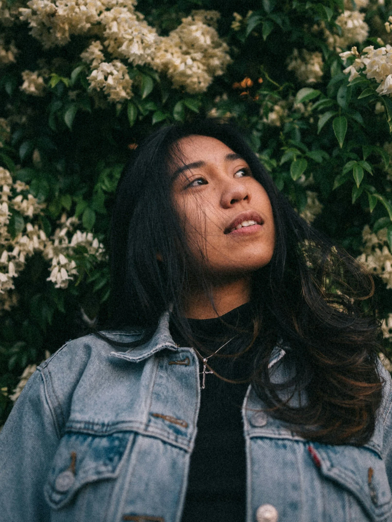 A young woman with long dark hair, wearing a denim jacket and black shirt, looking upward with a slight smile, standing in front of a bush with white flowers.