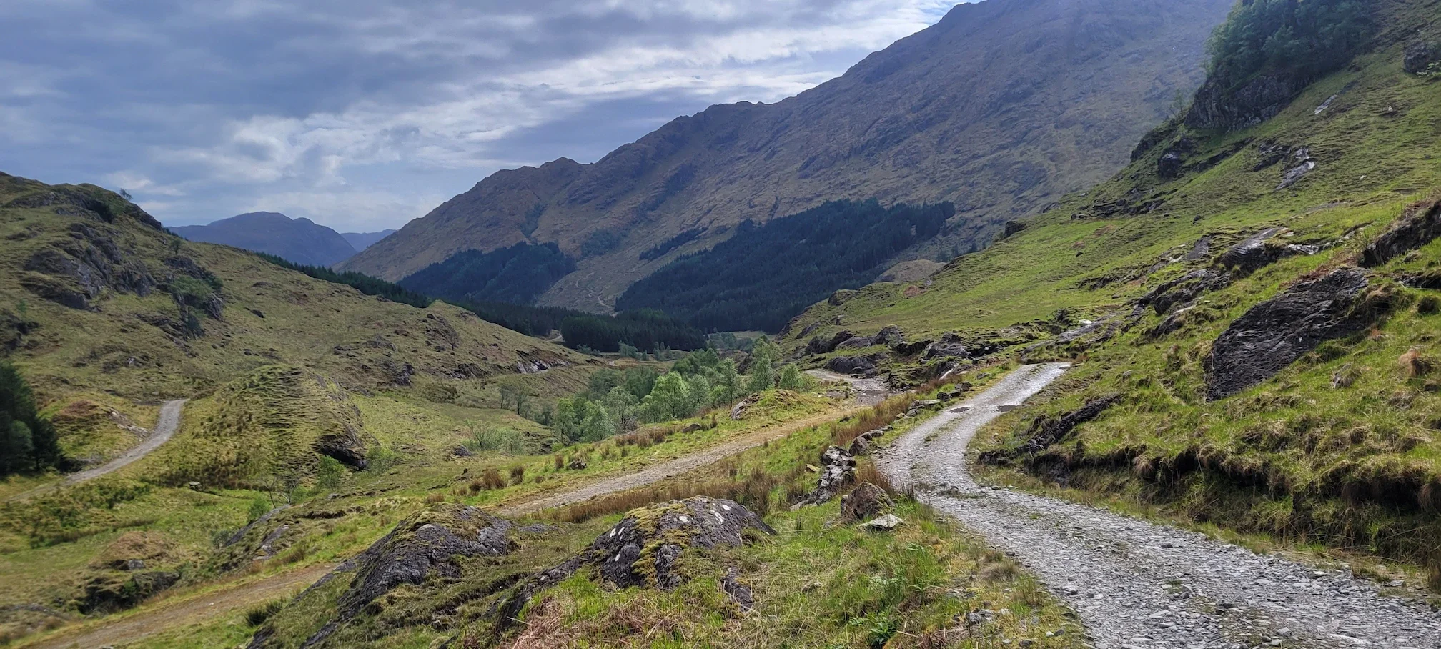 A dirt and gravel mountain trail winding through green hills and valleys with tall mountains in the background under a cloudy sky.
